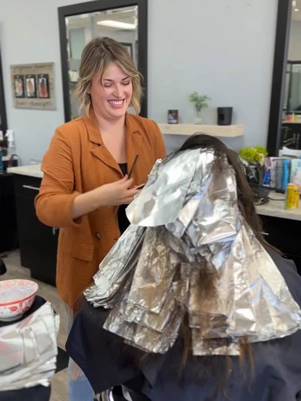 A hairstylist applies foils to a client's hair in a salon, with a cheerful and professional demeanor.