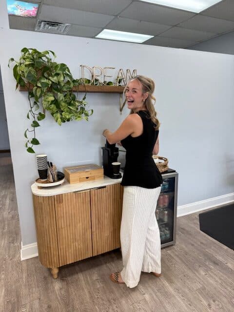 A woman with long hair stands by a coffee station, smiling while making coffee, surrounded by decorative plants and a cozy interior.